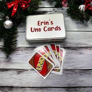 May include: A silver tin with the text "Erin's Uno Cards" in red letters. The tin is open and a deck of Uno cards is visible. The cards are red, yellow, green, and blue with the word "UNO" in white letters.