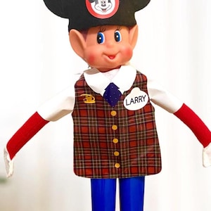 May include: A doll dressed as a character with a Mickey Mouse hat and a plaid vest. The doll has a white shirt, red sleeves, and blue pants. The name "LARRY" is on a white tag on the vest.