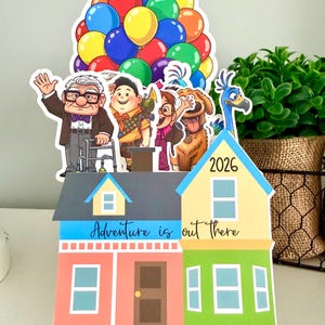 May include: A colorful paper cutout featuring characters and a house from the movie "Up." The house has the text "Adventure is out there" and the year "2026." A cluster of balloons tops the cutout.