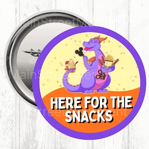 May include: A round button pin with a purple dragon illustration holding snacks. The dragon is on a yellow background with an orange banner that reads "HERE FOR THE SNACKS" in white text. The pin has a silver metal backing.