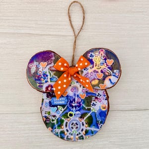 May include: A wooden Mickey Mouse-shaped ornament with a map design. The ornament features a bright orange polka-dot bow and a twine loop for hanging. The map design includes the text "Liberty Square".