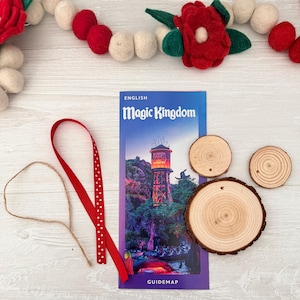 May include: A Magic Kingdom guidemap with a water tower illustration, three wooden coasters, a red ribbon with white polka dots, and a twine string. A felt garland with red and white balls and red flowers is in the background.