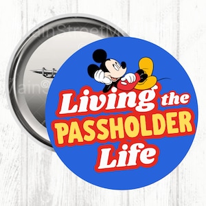 Op de afbeelding: Een ronde button met een blauwe achtergrond met Mickey Mouse en de tekst "Living the Passholder Life" in rood en geel. De button heeft een zilveren metalen rand en een speld.