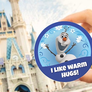 May include: A blue and white round button with a cartoon image of Olaf from Frozen. Olaf is smiling and has his arms outstretched. The text on the button reads "I like warm hugs!"