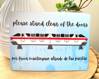 Könnte beinhalten: Ein blaues und weißes Schild mit einer roten und weißen Monorail-Illustration. Das Schild lautet "Please stand clear of the doors" auf Englisch und "Por favor mantengase alejado de las puertas" auf Spanisch.