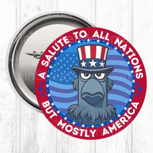 Op de afbeelding: Een ronde pin-back button met een cartoonadelaar die een rood-wit-blauw gestreepte hoge hoed met sterren draagt. De achtergrond is een blauw vlagontwerp. De rode randtekst luidt, A SALUTE TO ALL NATIONS BUT MOSTLY AMERICA.