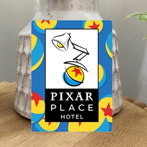 May include: A rectangular blue card with a Pixar theme. The card features the Pixar lamp and ball logo, with the words "PIXAR PLACE HOTEL" in a black rectangle. The background has a pattern of yellow stars and blue and yellow balls.