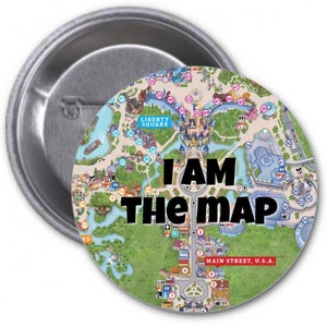 Puede incluir: Un botón redondo con un mapa de un parque de atracciones. El mapa está rodeado por un círculo negro con el texto "I AM THE MAP" en letras blancas. El mapa muestra las diferentes áreas del parque, incluyendo la calle principal, Liberty Square y el castillo.