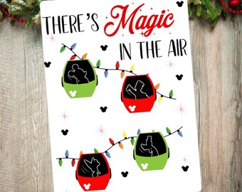 Könnte beinhalten: Ein weißes Schild mit dem Text "THERE'S Magic IN THE AIR" in Schwarz und Rot. Das Schild zeigt vier farbige Gondeln mit Charakter-Silhouetten, die mit Lichterketten und Mickey Mouse-Symbolen geschmückt sind.