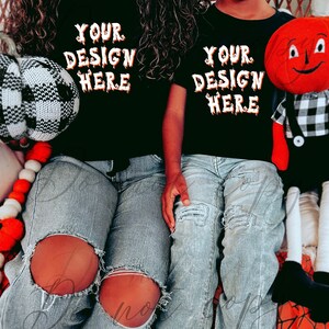 May include: Two people wearing black t-shirts with the text "Your Design Here" in orange dripping letters. The shirts are worn over blue jeans with distressed holes. The person on the right is holding a red plush toy with a black and white checkered pattern.
