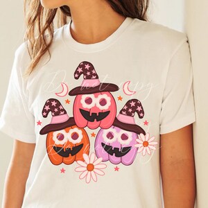 May include: Three cartoon pumpkins wearing witch hats with pink and brown accents. The pumpkins are orange, pink, and purple. They are surrounded by pink and white flowers and stars.