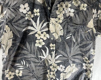 May include: A short-sleeved button-down shirt with a tropical floral pattern. The shirt features a dark gray background with white and light gray leaves and flowers. The shirt has a collar and a pocket on the left side.