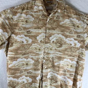 Puede incluir: Una camisa de manga corta con botones de color marrón y blanco con un estampado tropical de palmeras, islas y nubes. La camisa tiene un cuello blanco y un cierre de botones en la parte delantera.