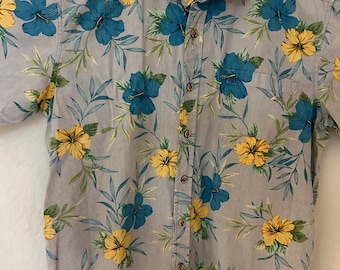 May include: A short-sleeved button-down shirt with a light gray background and a floral pattern of blue and yellow hibiscus flowers and green leaves. The shirt is hanging on a white hanger.