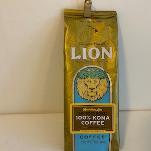 Pu&ograve; includere: Un sacchetto dorato di caff&egrave; Hawaiian Lion 24K Gold Roast 100% Kona, in grani interi, tostatura media-leggera. Il sacchetto presenta un leone con una ghirlanda di fiori e il testo "Honolulu, Hawaii" e "Proudly Roasting Since 1964". Il sacchetto pesa 7 once (198 g).