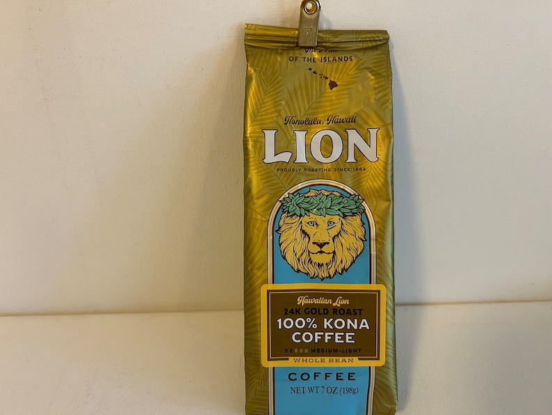 Pu&ograve; includere: Un sacchetto dorato di caff&egrave; Hawaiian Lion 24K Gold Roast 100% Kona, in grani interi, tostatura media-leggera. Il sacchetto presenta un leone con una ghirlanda di fiori e il testo "Honolulu, Hawaii" e "Proudly Roasting Since 1964". Il sacchetto pesa 7 once (198 g).