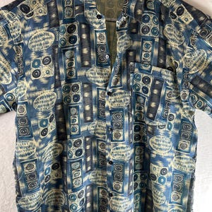 Puede incluir: Una camisa de manga corta con botones azul y beige con un patrón geométrico. La camisa tiene cuello y un bolsillo en el lado izquierdo.