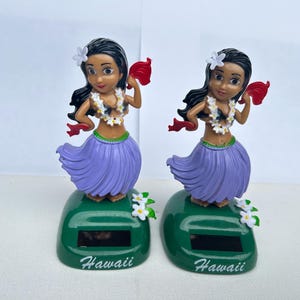 May include: Two solar-powered hula dancer figurines. Each figurine has a dark-haired woman wearing a white flower in her hair, a lei, a purple skirt, and holding a red fan. The figurines are on green bases with the word "Hawaii".