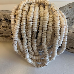 May include: A collection of off-white shell necklaces. The necklaces are made of small, flat, circular shells strung together. The shells have a natural, slightly irregular shape and texture. The necklaces are arranged in a tangled, layered bundle.