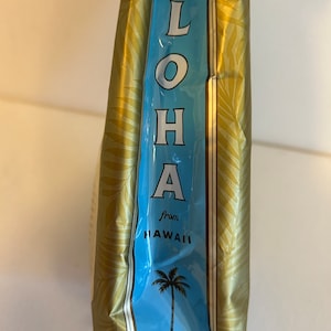 Pu&ograve; includere: Una borsa dorata con un'etichetta blu che dice "ALOHA from HAWAII" e una grafica di una palma.