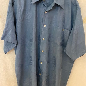 Puede incluir: Una camisa azul de manga corta con botones y estampado tropical. La camisa tiene un bolsillo en el lado izquierdo y el nombre de la marca "Blackpoint" es visible en el cuello.