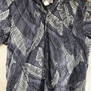 May include: A button-down shirt with a tropical leaf pattern in shades of gray and white. The shirt has a collar and short sleeves. The brand tag is visible at the collar. The shirt is hanging on a white hanger.