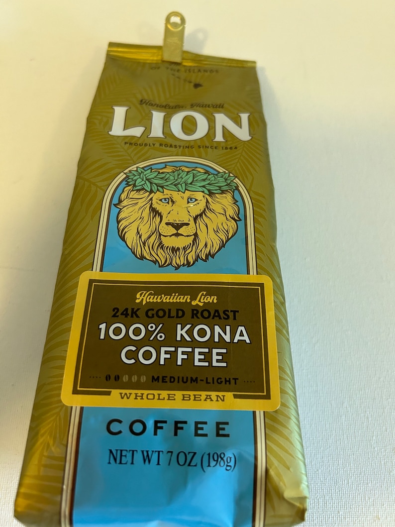 Pu&ograve; includere: Un sacchetto dorato di caff&egrave; Hawaiian Lion 24K Gold Roast 100% Kona, in grani interi, tostatura media-leggera. Il sacchetto presenta un leone con una corona di foglie e il testo "Hawaiian Lion 24K Gold Roast 100% Kona Coffee" in un riquadro giallo. Il sacchetto riporta anche il testo "Whole Bean" e "Net Wt 7 oz (198g)".
