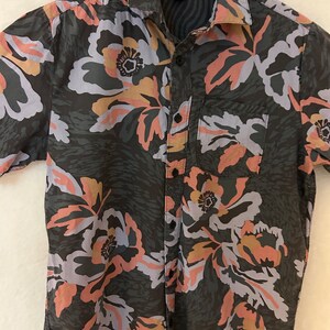 May include: A short-sleeved button-down shirt with a floral print in shades of pink, blue, and brown. The shirt has a pocket on the left side and a button-up collar.