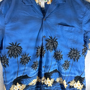 Może przedstawiać: Niebieska koszula z krótkim rękawem zapinana na guziki z nadrukiem palm i oceanu. Koszula ma kieszeń po lewej stronie i metkę z napisem "KY'S Made in Hawaii U.S.A."