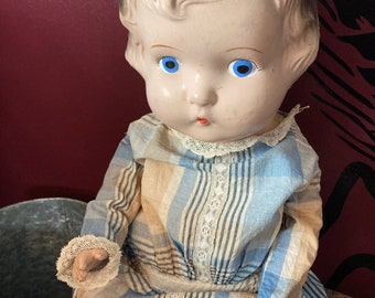 May include: Vintage doll with a painted face, blue eyes, and blonde hair. The doll wears a blue and tan plaid dress with lace trim. The doll's arms are visible, and the hands are painted.
