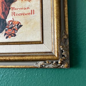 May include: A framed print of a painting by Norman Rockwell. The painting depicts a man and a woman sitting on a bench, with the man holding a book. The frame is gold with a decorative border.