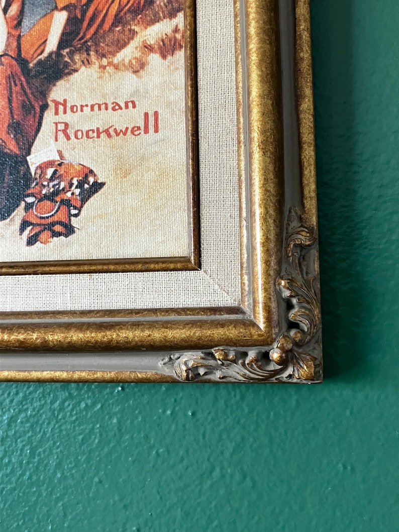 May include: A framed print of a painting by Norman Rockwell. The painting depicts a man and a woman sitting on a bench, with the man holding a book. The frame is gold with a decorative border.