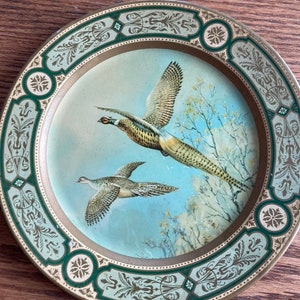 May include: A decorative round metal plate with a gold and green border. The center of the plate features a painting of two pheasants in flight against a blue sky with white branches.