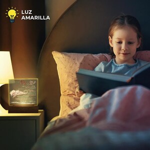 Peut inclure: Une fille lit un livre au lit. Un petit cube en verre lumineux avec le mot "Mama" et un c&oelig;ur est sur une table de chevet &agrave; c&ocirc;t&eacute; d'une lampe. La lumi&egrave;re du cube est jaune chaud.