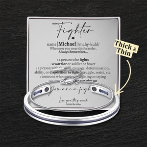 May include: A silver bracelet set with a blue cord and a silver charm that reads "You are a fighter". The bracelet comes with a card that reads "Fighter" and defines the word "fighter" as a person who fights, a warrior or soldier or boxer, a person with courage, determination, ability, or disposition to fight, struggle, resist, etc., someone who continues fighting or trying. The card also includes the name "Michael" and the phrase "Love you this much".