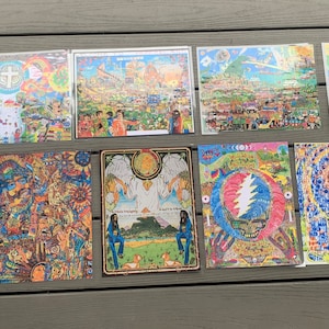 May include: A collection of nine colorful psychedelic posters featuring various designs, including a cross, a cityscape, a skull, and a winged figure. The posters are framed and displayed on a wooden surface.