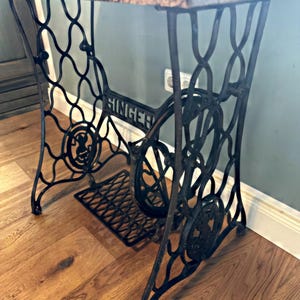 May include: A black cast iron Singer sewing machine base featuring an intricate, curved metal frame, a central flywheel, and a foot pedal. The base supports a thick, textured wooden tabletop with a natural grain finish and visible wood knots.