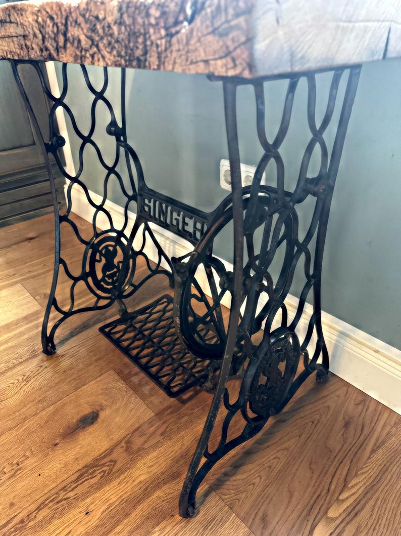 May include: A black cast iron Singer sewing machine base featuring an intricate, curved metal frame, a central flywheel, and a foot pedal. The base supports a thick, textured wooden tabletop with a natural grain finish and visible wood knots.