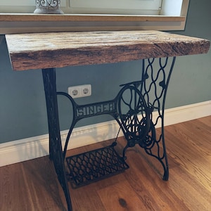 May include: A table featuring a thick, rectangular, weathered brown wooden top supported by a black cast iron base from a vintage Singer sewing machine, including the ornate metal legs, a large side wheel, and a foot pedal mechanism.