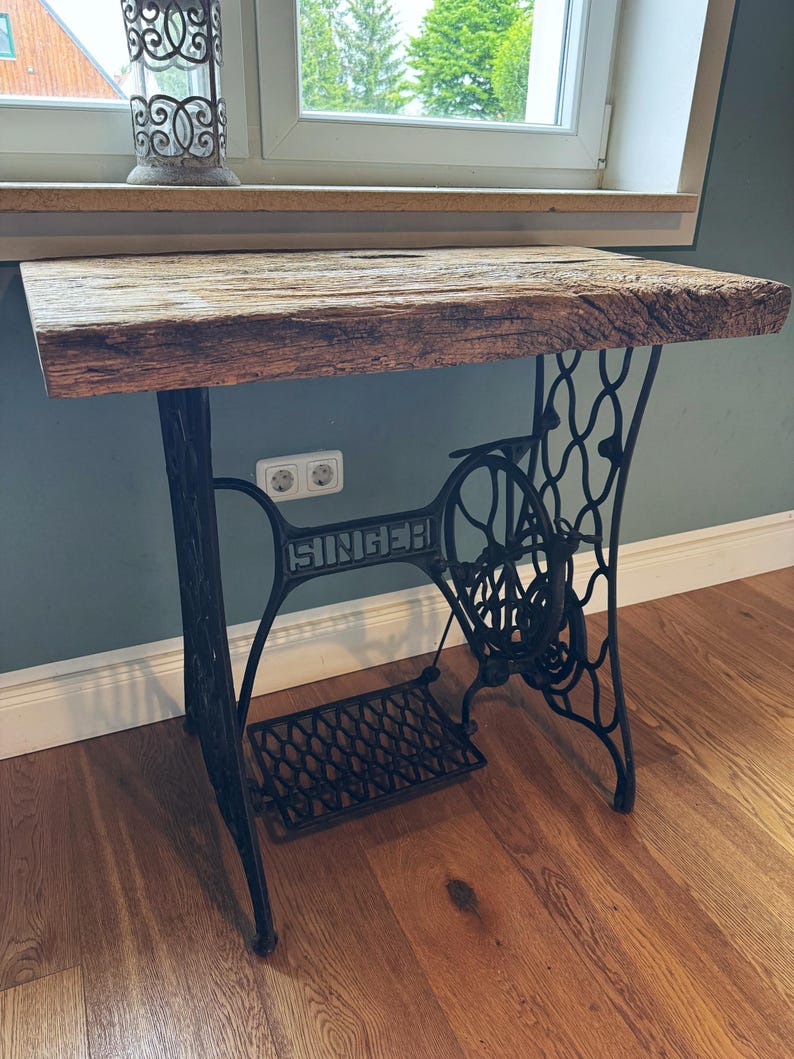 May include: A table featuring a thick, rectangular, weathered brown wooden top supported by a black cast iron base from a vintage Singer sewing machine, including the ornate metal legs, a large side wheel, and a foot pedal mechanism.
