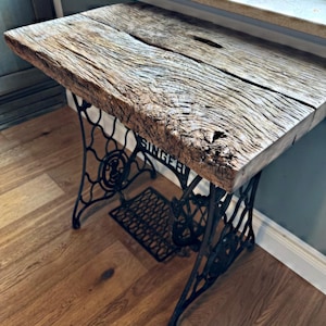 May include: A table featuring a thick, weathered brown wooden slab top with a prominent natural grain and a central fissure, supported by a black cast iron base with intricate scrollwork and the word SINGER embossed on the side frame.