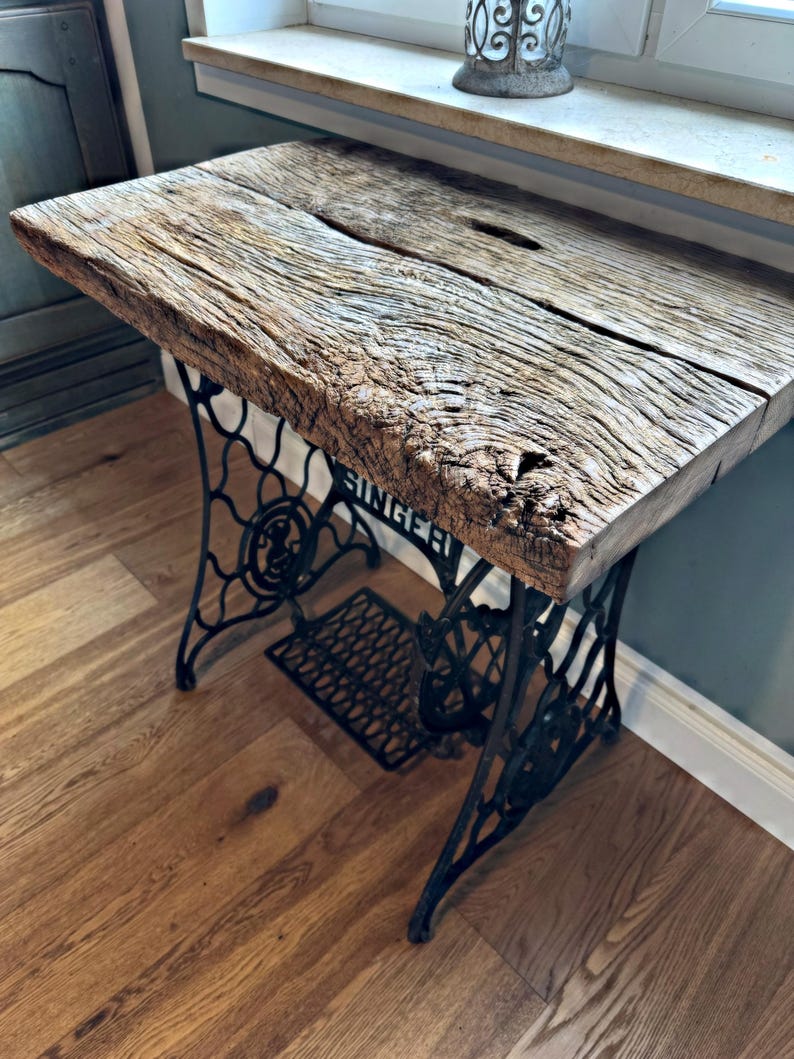 May include: A table featuring a thick, weathered brown wooden slab top with a prominent natural grain and a central fissure, supported by a black cast iron base with intricate scrollwork and the word SINGER embossed on the side frame.