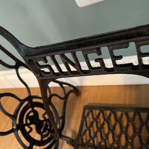 May include: A black cast iron Singer sewing machine base featuring an intricate, curved metal frame design with the brand name SINGER embossed in capital letters across the central horizontal support bar and a treadle wheel component.
