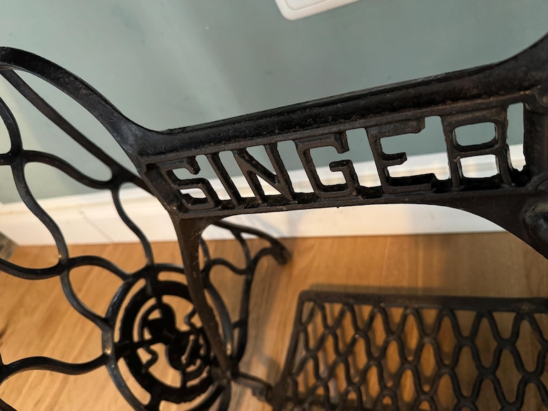 May include: A black cast iron Singer sewing machine base featuring an intricate, curved metal frame design with the brand name SINGER embossed in capital letters across the central horizontal support bar and a treadle wheel component.
