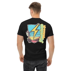 May include: Black t-shirt featuring a graphic of a yellow lightning bolt over a stylized landscape with the text "American Tradesman Co." in a retro font. The design includes a blue, yellow, and pink color scheme.