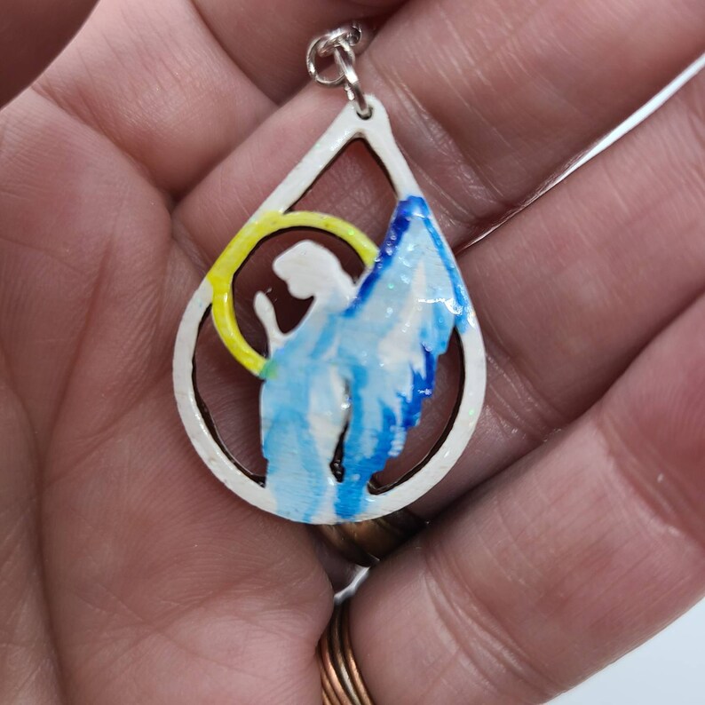 May include: A white teardrop-shaped pendant with a cutout design of an angel with wings. The angel is painted in shades of blue and white, with a yellow halo.
