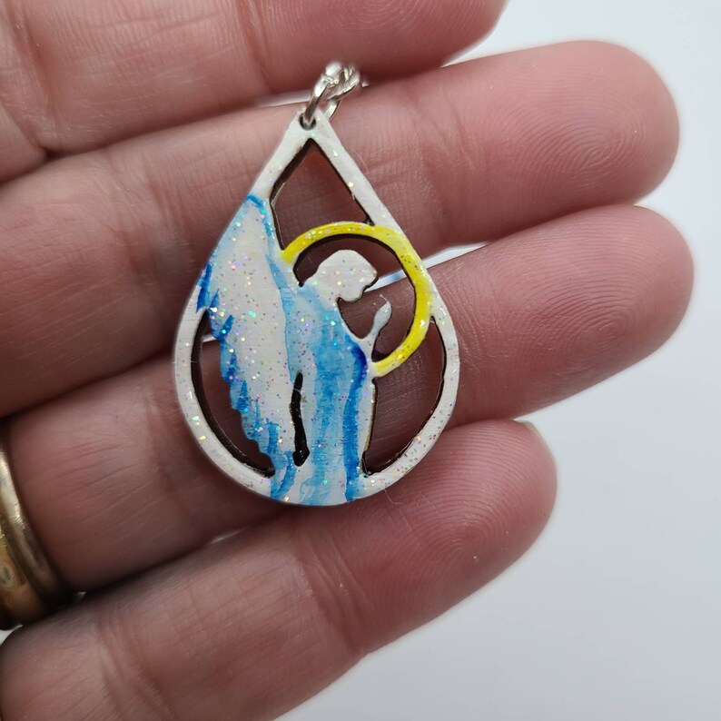 May include: A white teardrop-shaped pendant with a blue and white angel silhouette inside. The angel has a halo with a yellow outline. The pendant is made of wood and has a glitter finish.