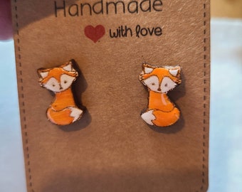 Könnte beinhalten: Ein Paar Holzohrringe in Form von Füchsen mit orangefarbenem Fell und weißen Schwänzen. Die Ohrringe befinden sich auf einer braunen Karte mit dem Text "Handmade with love".