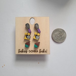 May include: A set of wooden earrings on a light wood backing. The earrings depict baking-related items: whisks with purple handles, yellow stand mixers, and cupcakes with teal frosting. The text "Bakers GONNA Bake!" is engraved on the wood.