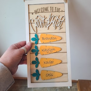 May include: Wooden sign with a white frame and the words "WELCOME TO THE Carrot Patch" in a cursive font. Below are four orange carrot shapes with names: Brandon, Kaylyn, Xavier, and Otis. The carrots have green leaves.